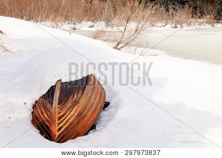 Old Wooden Destroyed Flooded Fishing Boat Lie In Snow On Shore Of Ice-covered Frozen River In Winter