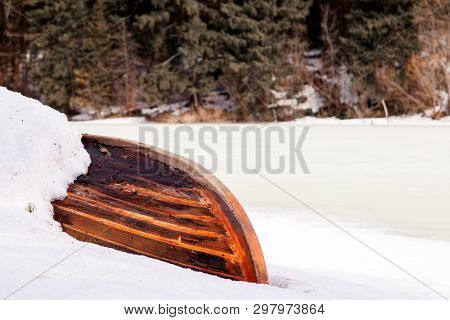 Old Wooden Destroyed Flooded Fishing Boat Lie In Snow On Shore Of Ice-covered Frozen River In Winter