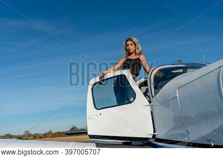 A gorgeous young blonde model poses outdoors with a single engine aircraft at a local airport