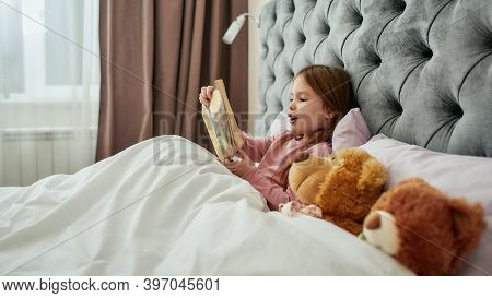 A Little Cute Girl Holding Her Book Closely And Enjoying Reading Aloud While Lying In A Big Bed With
