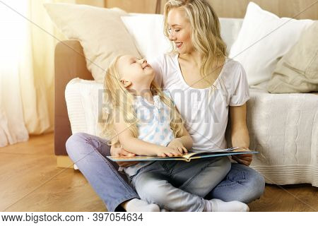 Happy Family. Blonde Young Mother Reading A Book To Her Cute Daughter While Sitting At Wooden Floor 