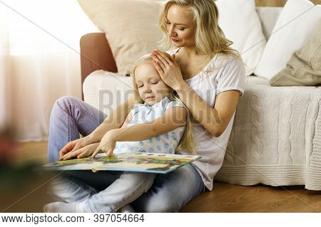 Happy Family. Blonde Young Mother Reading A Book To Her Cute Daughter While Sitting At Wooden Floor 