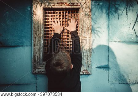Window With Bars. A Hand Gripped An Old Metal Grate. The Boy Is Holding On To The Bars. The Child Cl