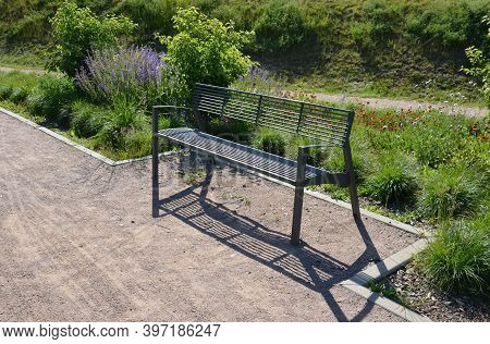 Gray Metal Park Bench With Transparent Design Of Metal Wires Cross Bars Path In The Park Flowerbed I