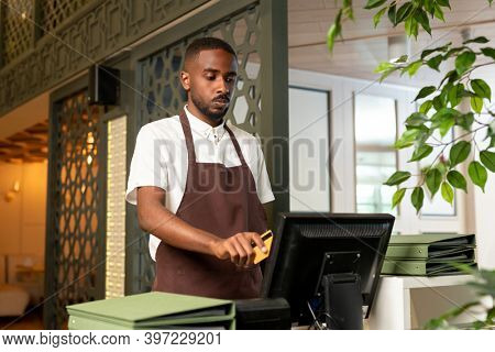 Young waiter or cashier in shirt and apron standing in front of computer and registering payment of client for the ordered food and drink