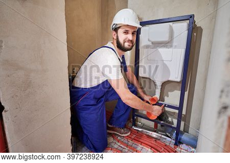 Handsome Bearded Man In Work Overalls Installing Concealed Toilet Frame In Bathroom. Cheerful Male W