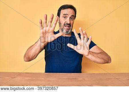 Middle age hispanic man wearing casual clothes sitting on the table afraid and terrified with fear expression stop gesture with hands, shouting in shock. panic concept. 