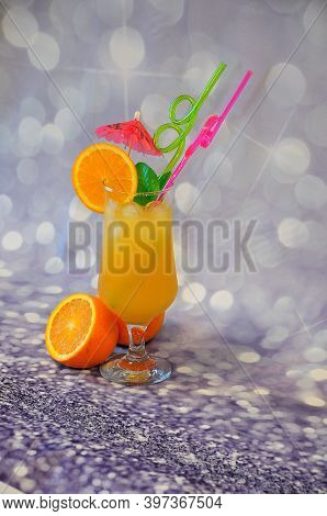 A Glass Of Citrus Juice With Ice, Decorated With An Umbrella, Straws And Whole And Cut Orange Fruits