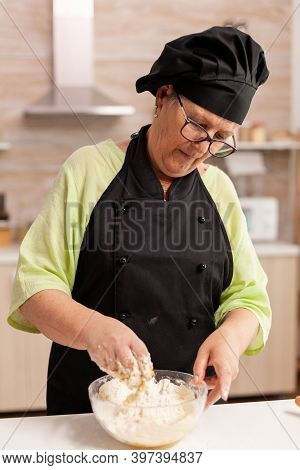 Old Pastery Chef Wearing Bonete And Apron With Hand In Wheat Flour With Eggs. Retired Elderly Chef W