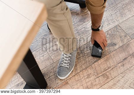 Cropped View Of Businessman Taking Smashed Cellphone From Wooden Floor With Blurred Table On Foregro
