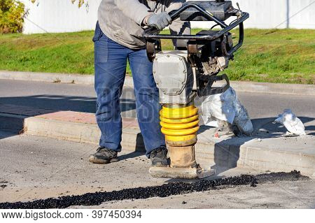 A Road Worker Uses A Petrol Vibratory Rammer To Repair Asphalt On The Roadway On A Bright Sunny Day.