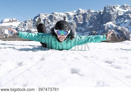A Teenage Girl In A Colorful Ski Suit, Helmet And Sunglasses Lies In The Snow And Smiles Happily Aga