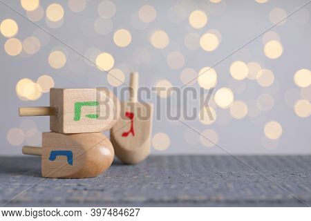 Hanukkah Traditional Dreidels On Grey Wooden Table Against Blurred Lights. Space For Text