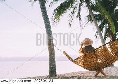 A Young Woman In Straw Hat Sitting In A Hammock Swinging Between A Palm Trees On The Overseas Island