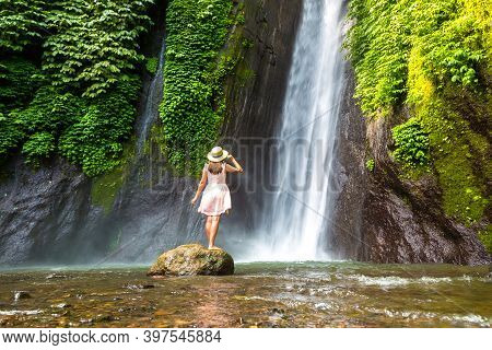 Woman Traveler At  Munduk Waterfall In Bali, Indonesia In A Sunny Day