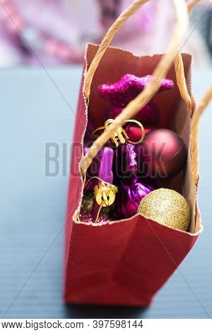 Red Paper Bag With Small Red, Violet And Golden Christmas Tree Toys On Grey Table. Rose Blurred Back