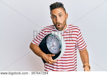 Handsome man with beard holding weight machine to balance weight loss scared and amazed with open mouth for surprise, disbelief face 