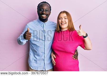 Young interracial couple expecting a baby, touching pregnant belly doing happy thumbs up gesture with hand. approving expression looking at the camera showing success. 