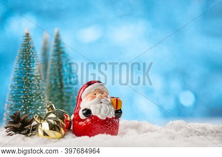Ceramic Figure Of Santa Claus, Fir Cones And Balls In The Snow Among The Christmas Trees On An Abstr