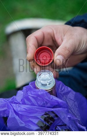 Man Opens A Bottle Of Alcoholic Drink Hidden In Violet Bag In The Park. Whisky Consumption Outdoors.