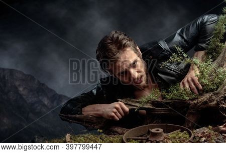 A Young Bearded Man In A Black Leather Jacket Climbs Out Of The Pit Holding On To A Snag. Expressive
