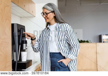 Happy white-haired mature woman smiling while using coffee maker indoors