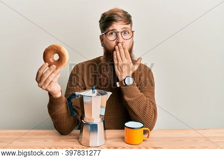 Young irish redhead man sitting on the table having breakfast covering mouth with hand, shocked and afraid for mistake. surprised expression 