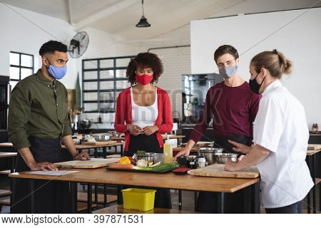 Female chef and divserse group wearing face masks in kitchen. trainee chefs standing around a table listening to female chef. hygiene in professional kitchen during coronavirus covid 19 pandemic.