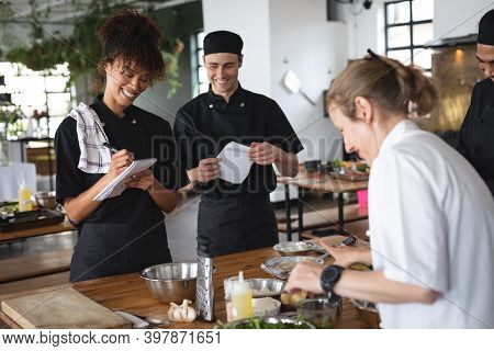 Female chef and divserse group in kitchen. trainee chefs standing around a table listening to female chef.