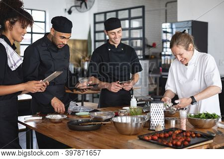 Female chef and divserse group in kitchen. trainee chefs using tablets and standing around a table listening to female chef.