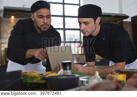 Two divserse male chefs in kitchen. two trainee chefs standing at a table and using tablet.