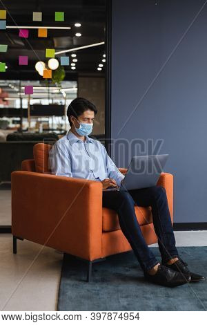 Caucasian businessman wearing face mask using laptop in creative office. social distancing in business office workplace during covid 19 coronavirus pandemic.