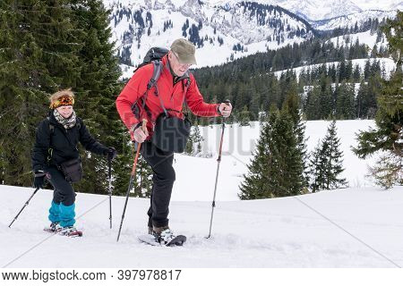 Senior Couple Is Snowshoe Hiking In Alpine Snow Winter Mountains. Allgau, Bavaria, Germany.