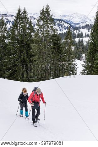 Senior Couple Is Snowshoe Hiking In Alpine Snow Winter Mountains. Allgau, Bavaria, Germany.