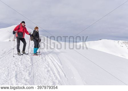 Senior Couple Is Snowshoe Hiking In Alpine Snow Winter Mountains. Allgau, Bavaria, Germany.