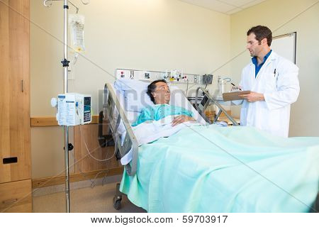 Young doctor looking at patient while writing notes on clipboard in hospital room