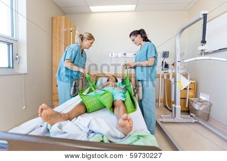 Female nurses preparing male patient before transferring him on sling lift on bed at hospital