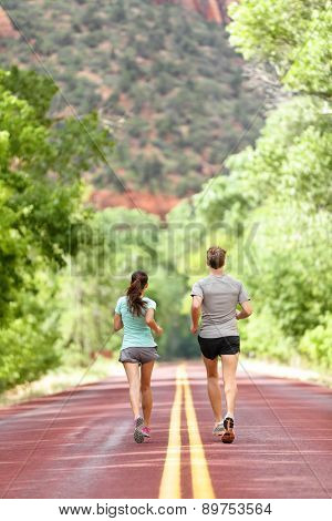 Runners running on road in nature away from camera. Couple, woman and man jogging for a run outside in amazing mountain landscape. Full body length rear view of back. Fitness and healthy lifestyle.