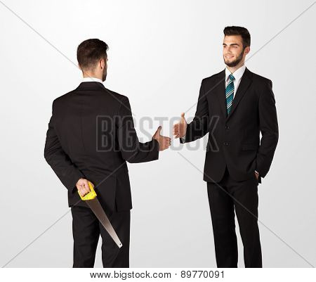 Two opposing businessman shake hands, one of them hiding a weapon behind his back