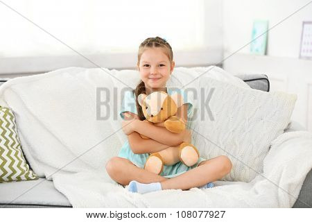 Beautiful little girl with teddy bear sitting on sofa, on home interior background