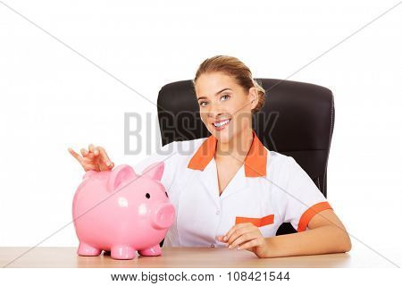 Young smile female doctor or nurse sitting behind the desk and holding a piggybank.