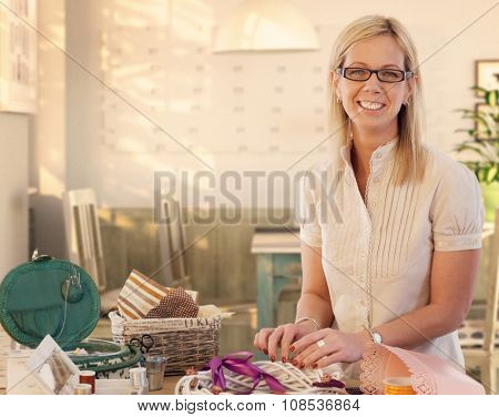 Happy casual caucasian blonde mid adult businesswoman making valentine day wreath at home. Smiling, wearing glasses, standing at table, do it yourself, gift, present. Looking at camera.