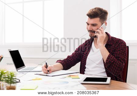 Important call. Young smiling businessman in casual talking on mobile phone in modern white office interior.