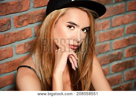 Cool trendy girl in baseball cap is leaning against a brick wall. Hipster style. Youth fashion.