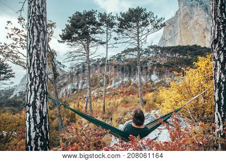 Back view of traveler man hiking and relaxing in hammock in autumn woods and looking at mountain hill. Cold weather, fall colors. Wanderlust concept.