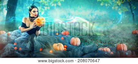 Halloween Witch with a carved Pumpkin and magic lights in a dark forest. Beautiful young surprised woman in witches costume holding pumpkin. Wide Halloween party wide angle art design