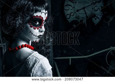 Little girl with santa muerte makeup for halloween is standing in left of the photo looking over her shoulder. Girl has black curly hair white dress and red bead