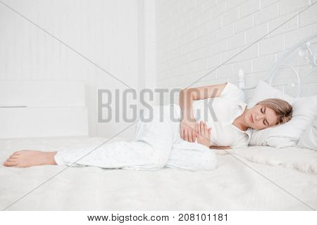 Blonde, young girl in white clothes lies on a bed with a pain in the stomach on a white background