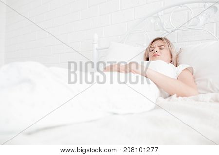 Blonde, young girl in white clothes lies on a bed with a pain in the stomach on a white background