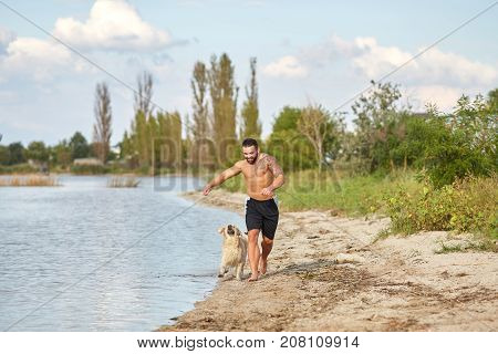 A young man with muscular body runs on the beach with his friend dog. An owner and dog walking on a nature background.